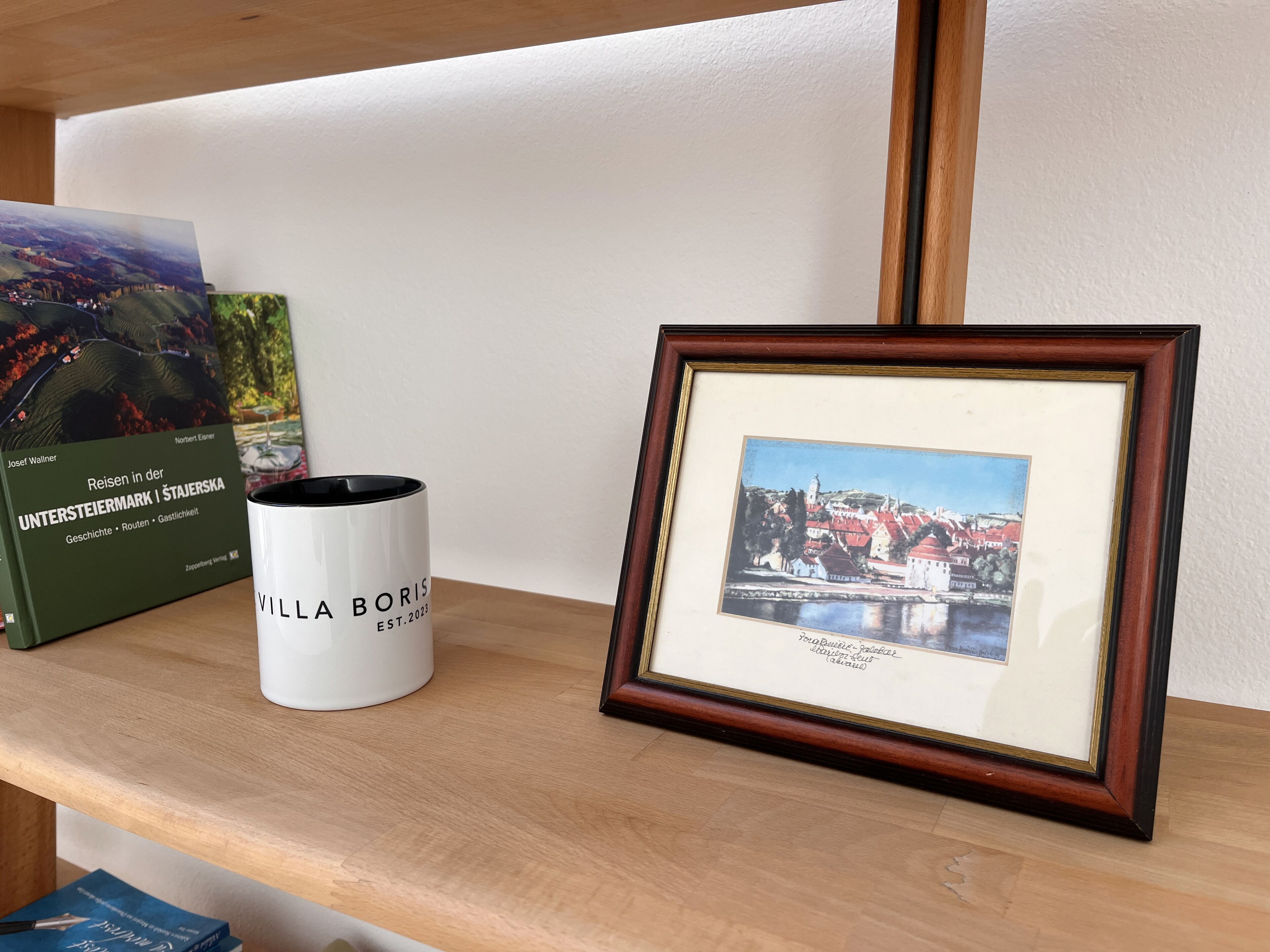 A framed picture of a town, a white mug, and two books on a shelf.