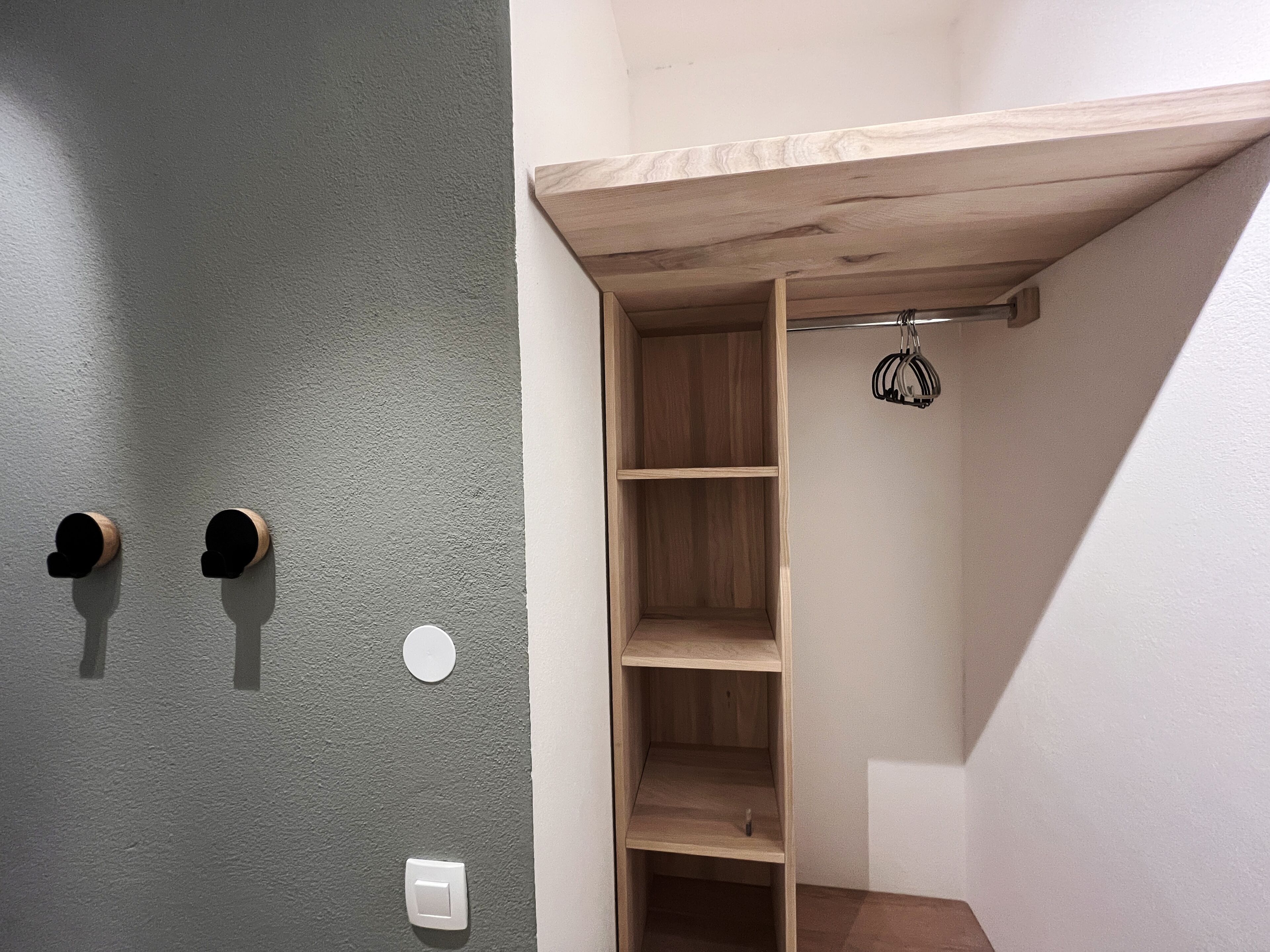 A corner of a room with a grey textured wall, a wooden shelf unit, and a hanging rack.