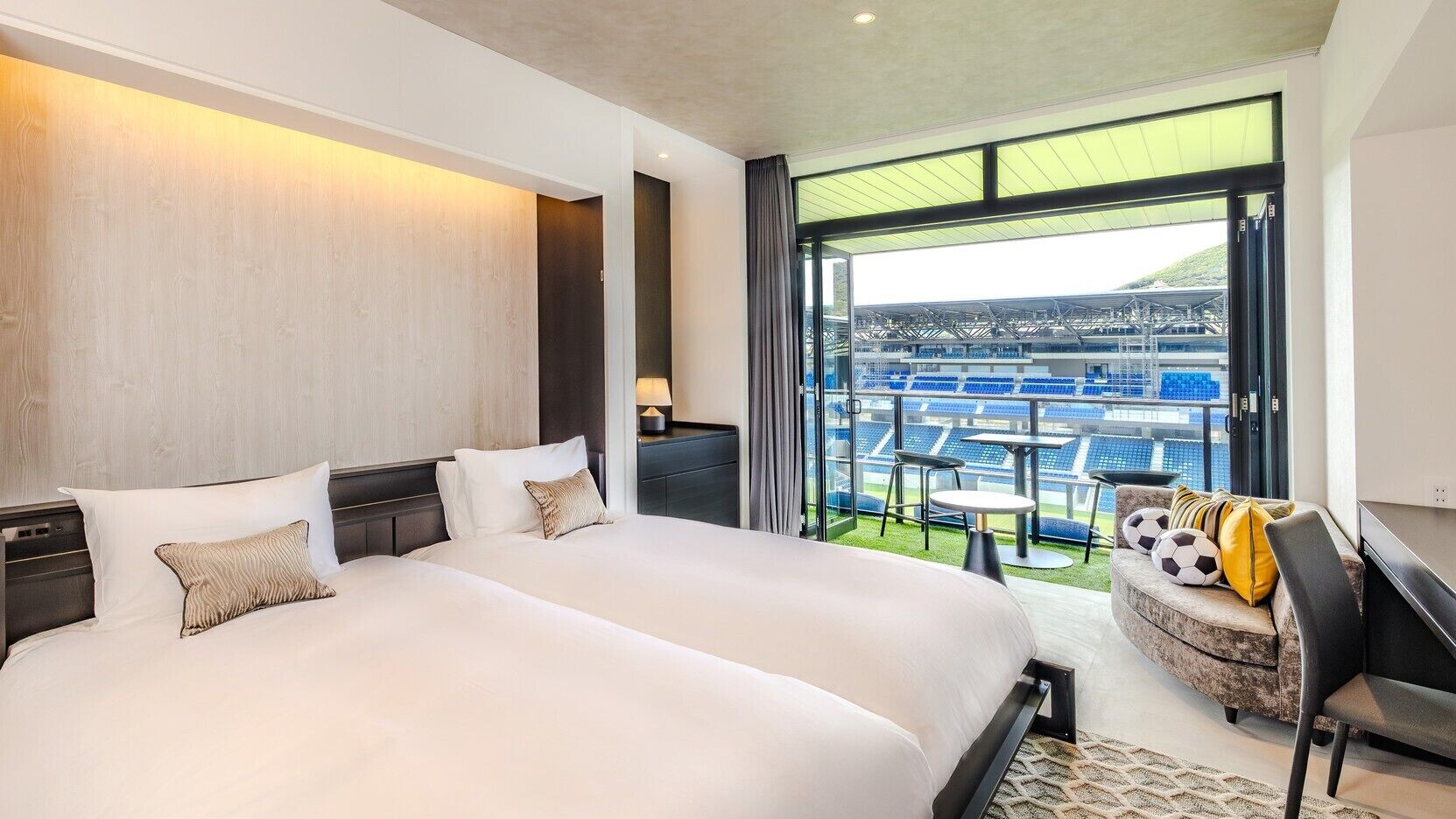 A modern hotel room with a large bed, a seating area with a view of a stadium, and a balcony with a view of the stadium.