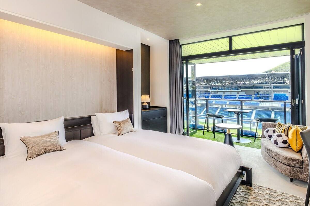 Una moderna camera d'albergo con un letto grande, un'area salotto con vista su uno stadio e un balcone anch'esso con vista sullo stadio.