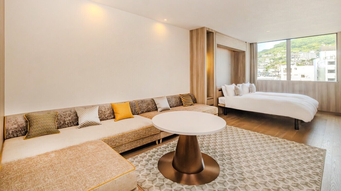 A hotel room with a sofa, a round table, a bed, and a large window.
