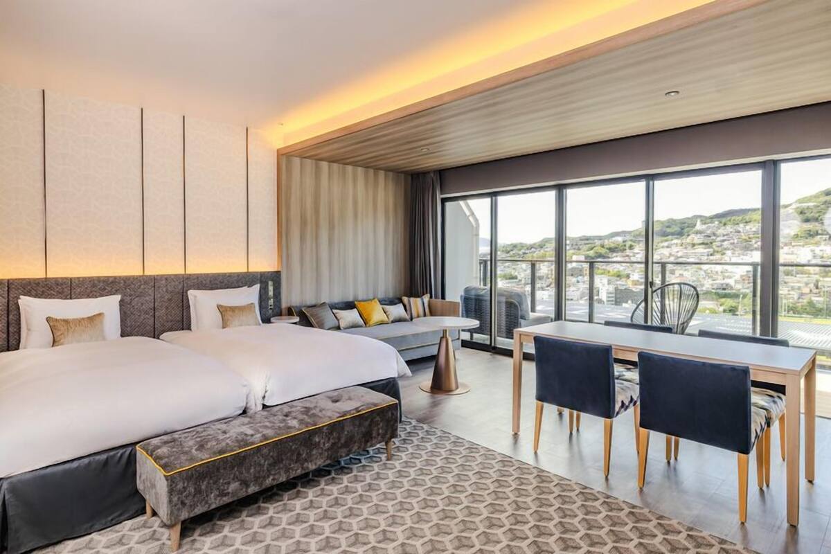 Una camera d'hotel moderna con un letto grande, un divano, un tavolo da pranzo con sedie e un balcone con vista sulla città.