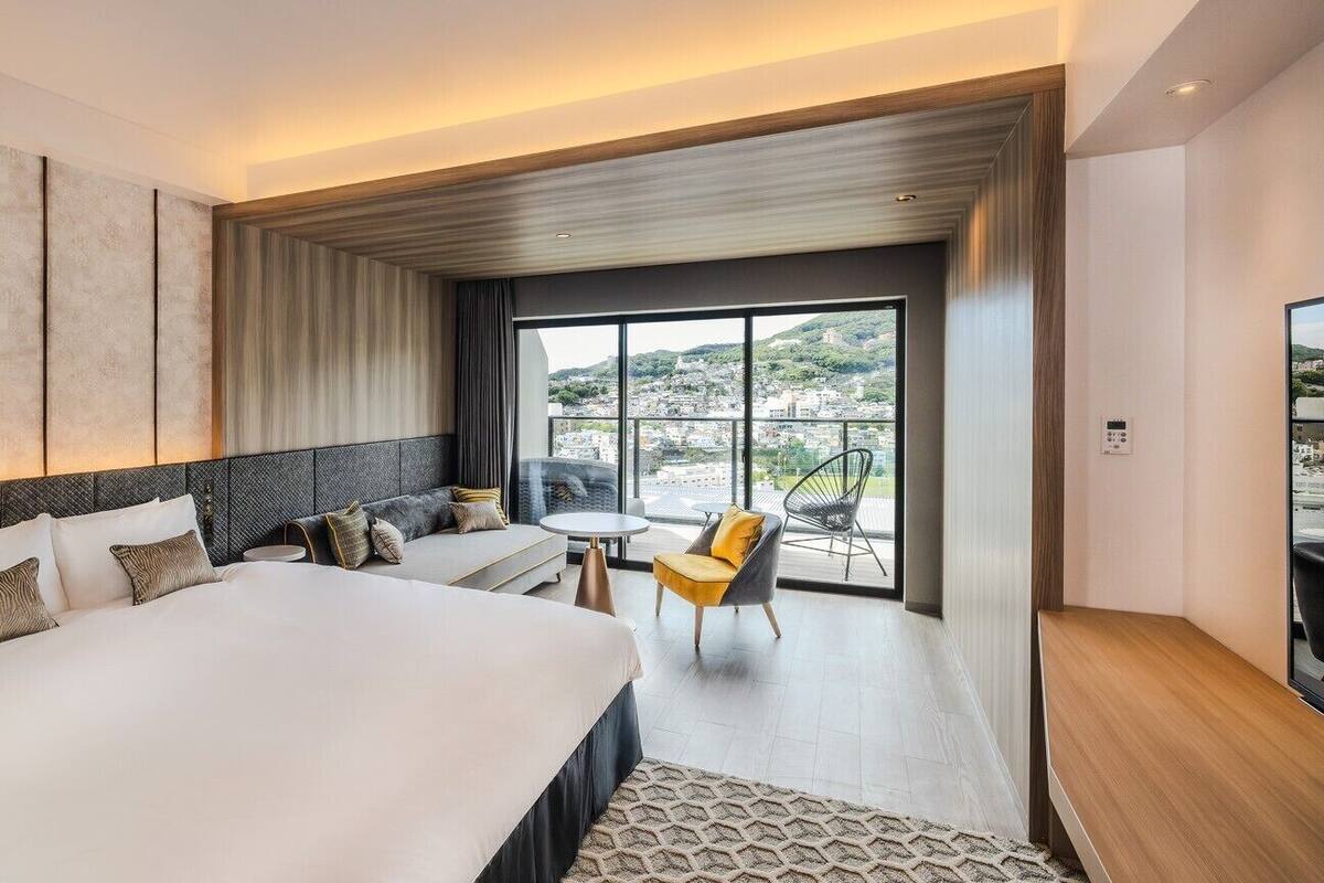 Una camera d'albergo con un letto grande, un divano, un tavolino, una sedia e un balcone con vista sulla città.