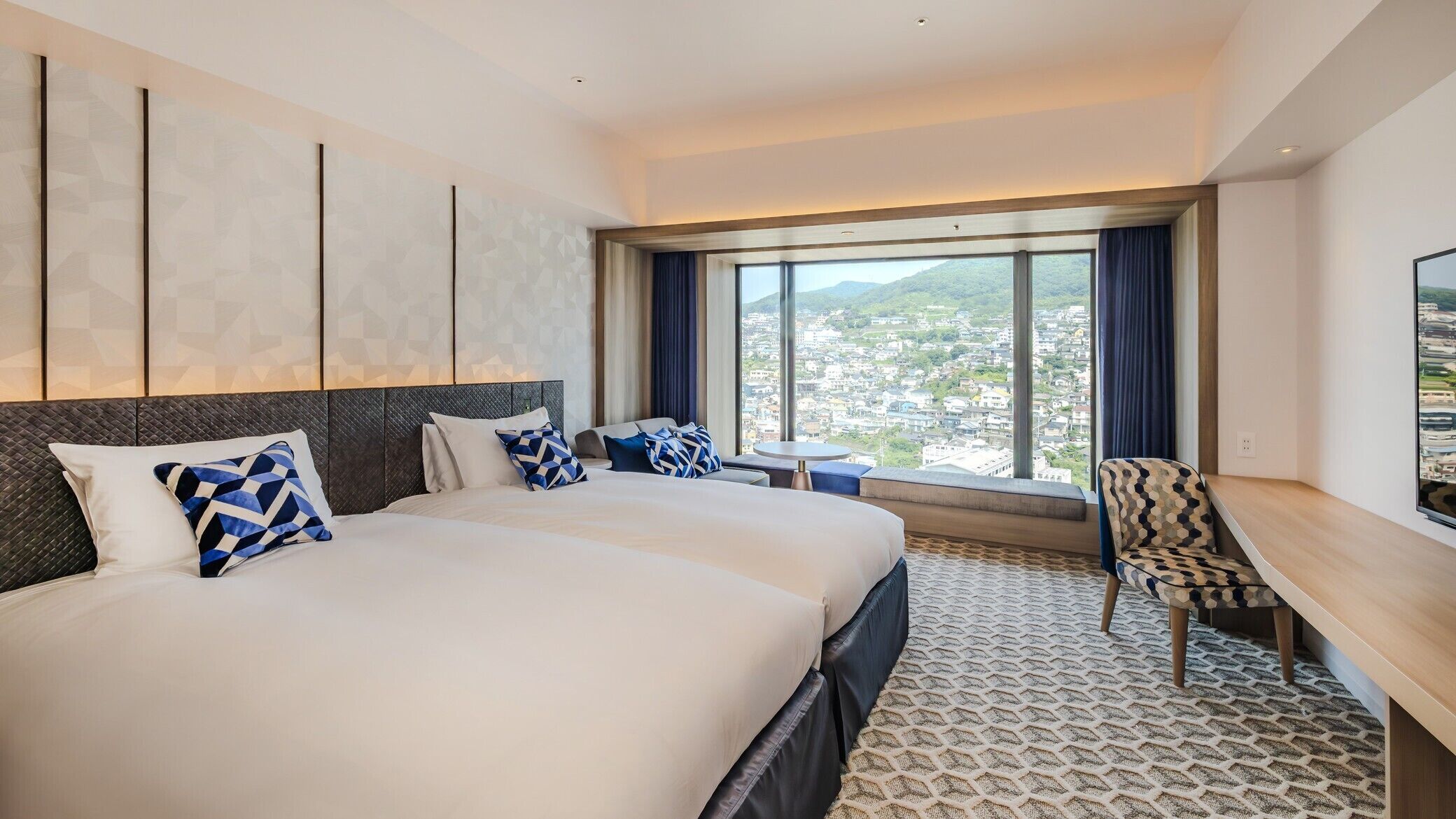 A hotel room with a large bed, a desk, a chair, and a large window with a city view.