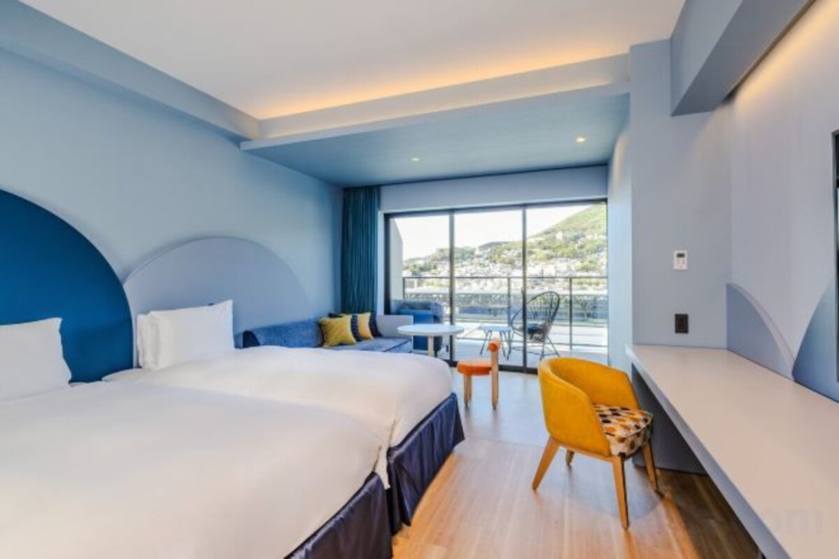 Habitación de hotel con una cama grande, zona de estar con mesa y sillas, y vistas al exterior.