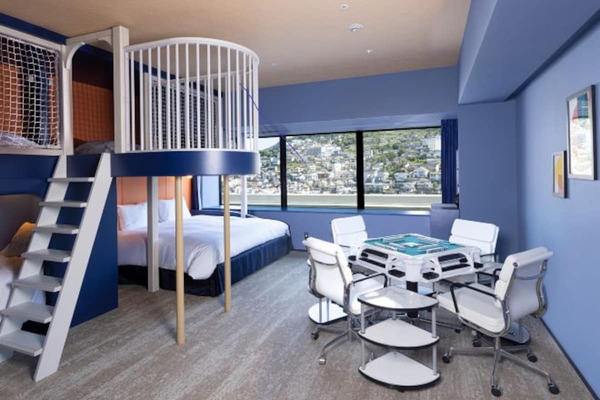 A room with a bunk bed, a desk with chairs, and a large window offering a city view.