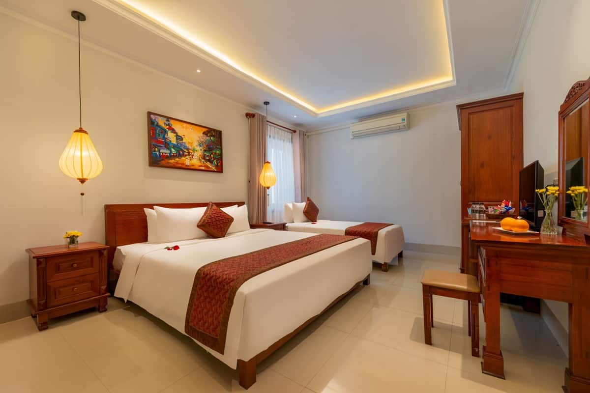 Une chambre d’hôtel avec un grand lit, un plus petit lit, un bureau et une chaise. Il y a un tableau au mur et une fenêtre avec des rideaux.