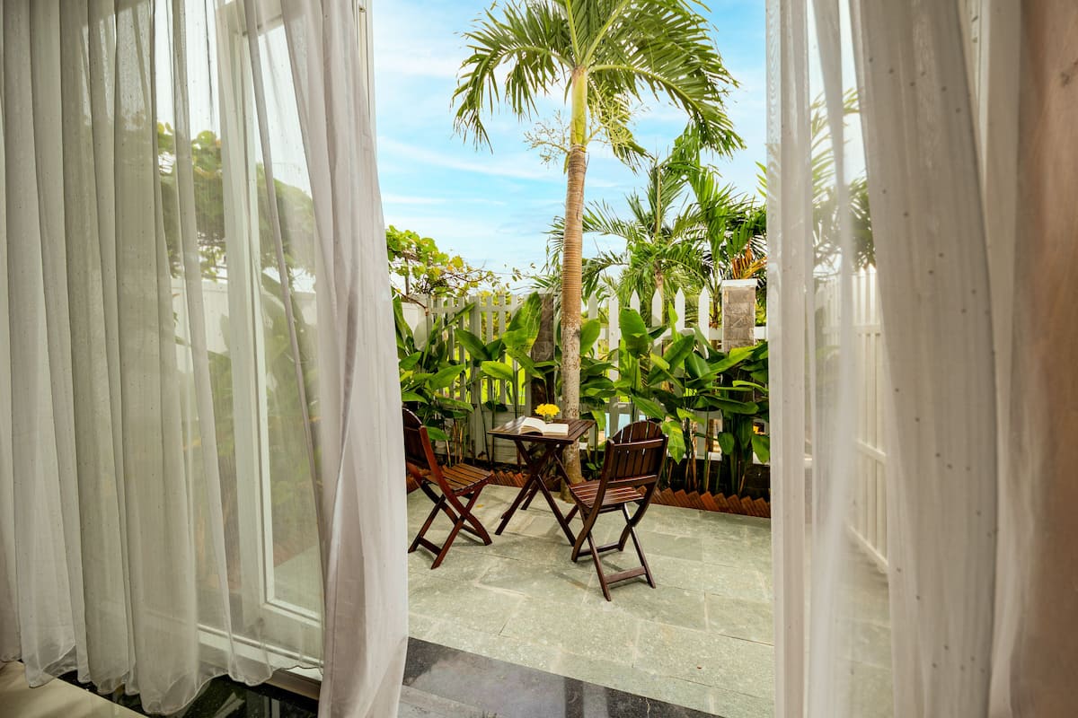 Une vue à travers des rideaux légers sur un espace patio avec une table et des chaises, entouré d’une végétation luxuriante et d’un grand palmier.