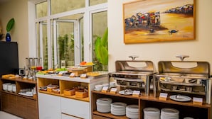 A buffet setup with multiple serving stations, a painting on the wall, and a view of the outdoors through large windows. - Iris De Villa Hoi An (Da Nang)
