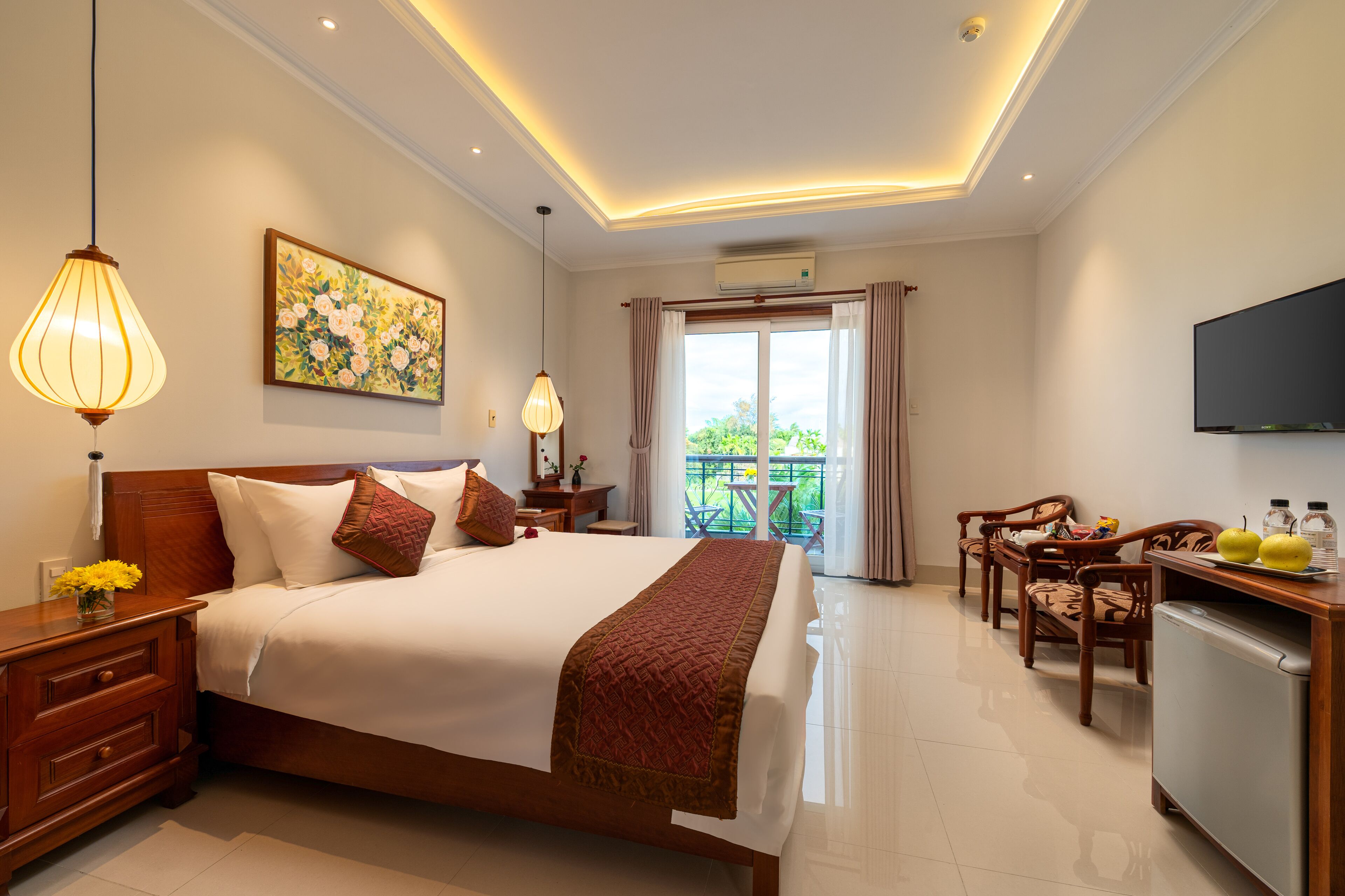 A hotel room with a large bed, a desk with a refrigerator, a dining area with chairs, a painting on the wall, and an air conditioning unit.