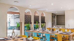 A restaurant with multiple tables and chairs, some with yellow and blue chairs, and others with wooden chairs. There are potted plants and a decorative screen with a geometric pattern. - SAPPHIRE BLISS HOTEL (Hyderabad)