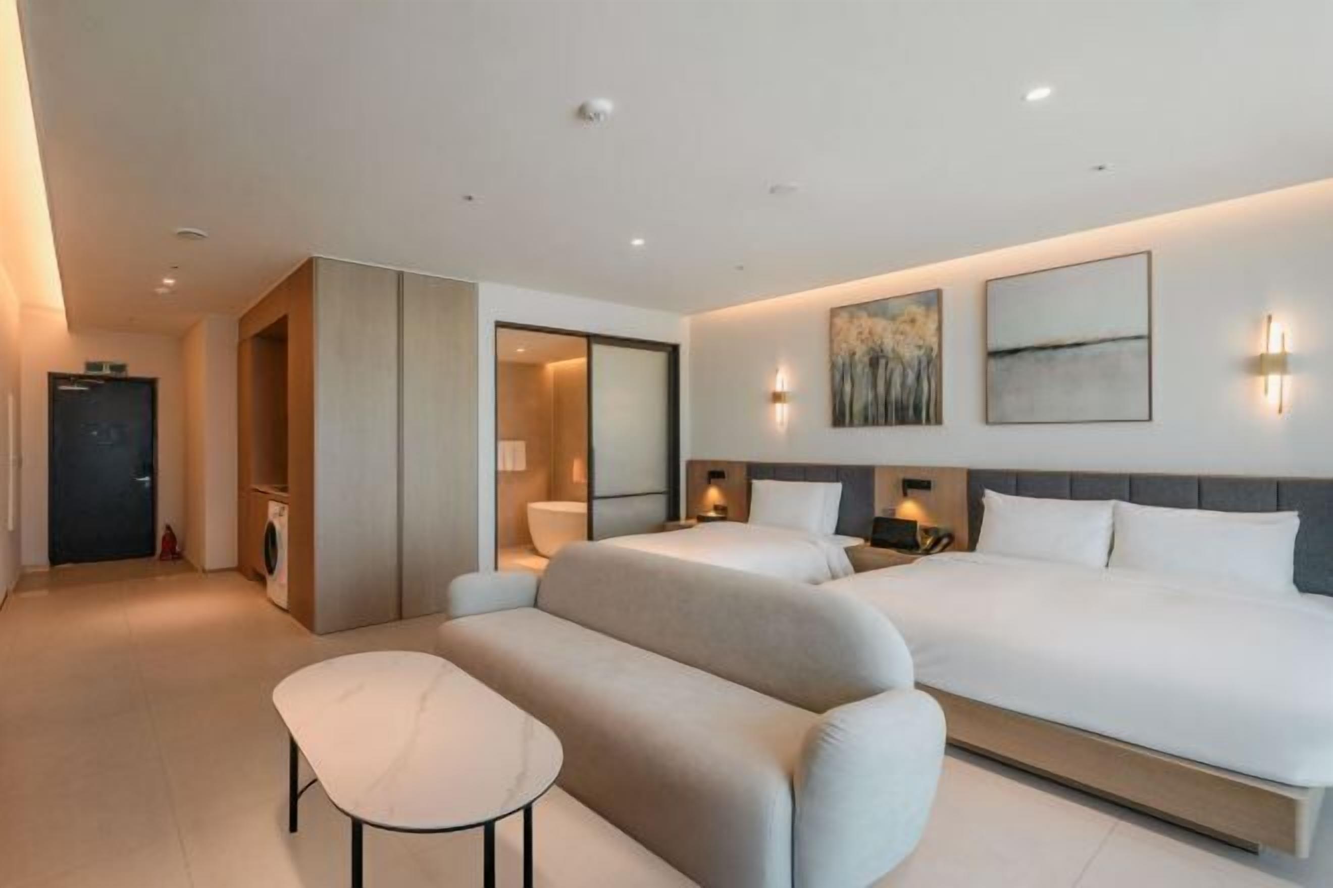 A modern hotel room with a large bed, a sofa, a small table, and a bathroom with a mirror and toilet.