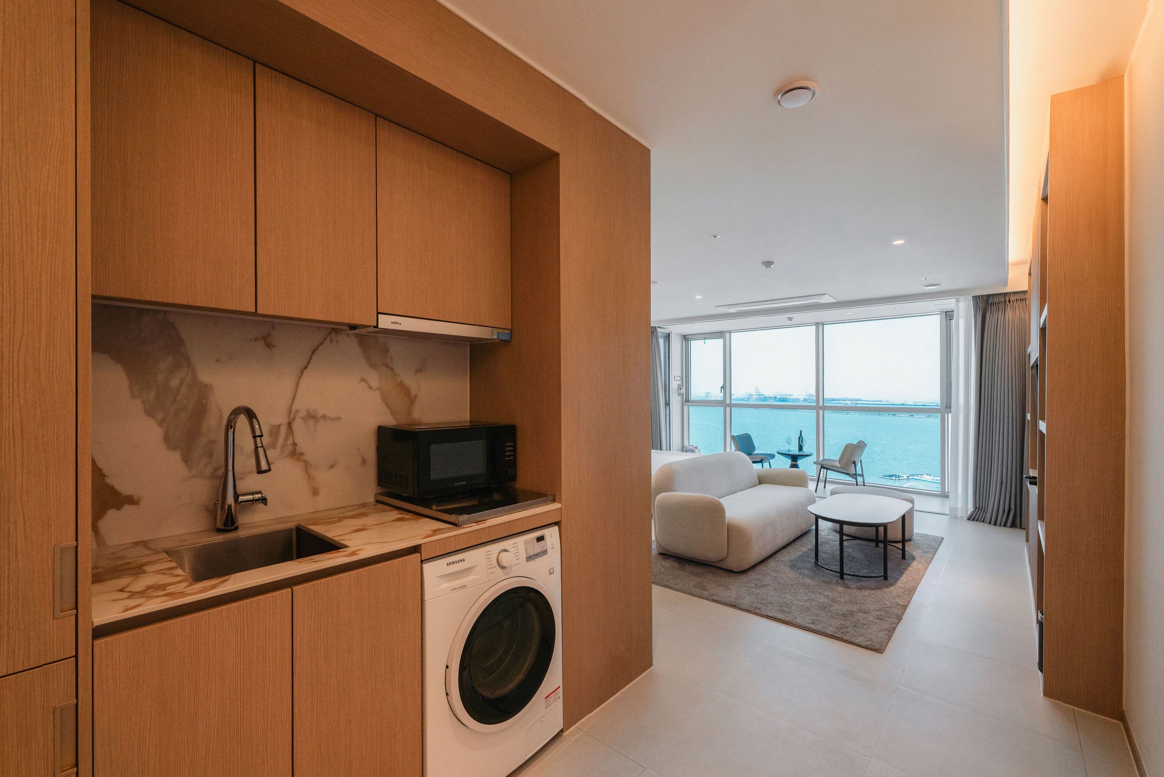 A modern kitchen with a washing machine, a sink, and a microwave. There is a living area with a sofa and a table, and a large window offering a view of the sea.