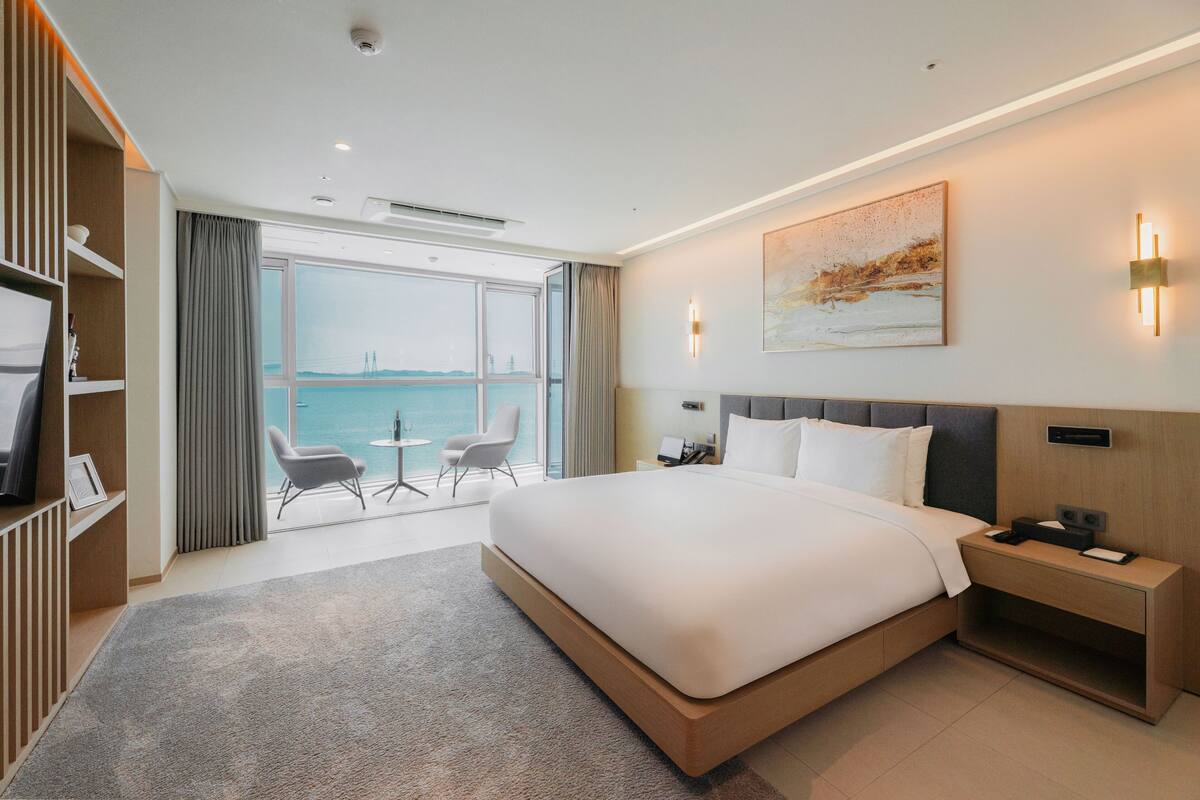 A modern hotel room with a large bed, bedside tables, a desk, and a view of the sea.