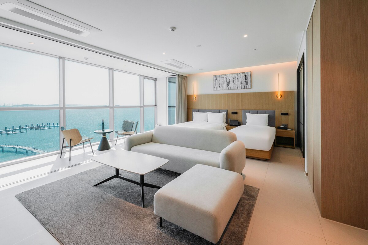 A modern hotel room with a large window overlooking a body of water, a bed, a sofa, a small table, and a chair.