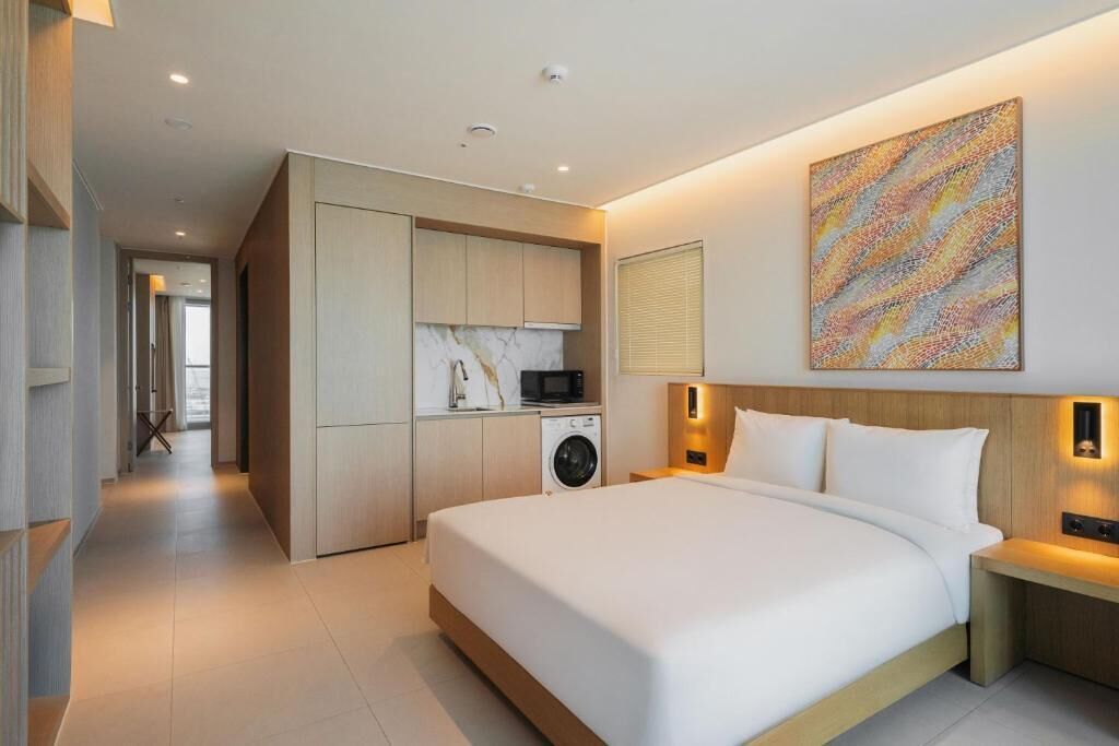 A modern hotel room with a large bed, built-in kitchen with appliances, and a view of the hallway.