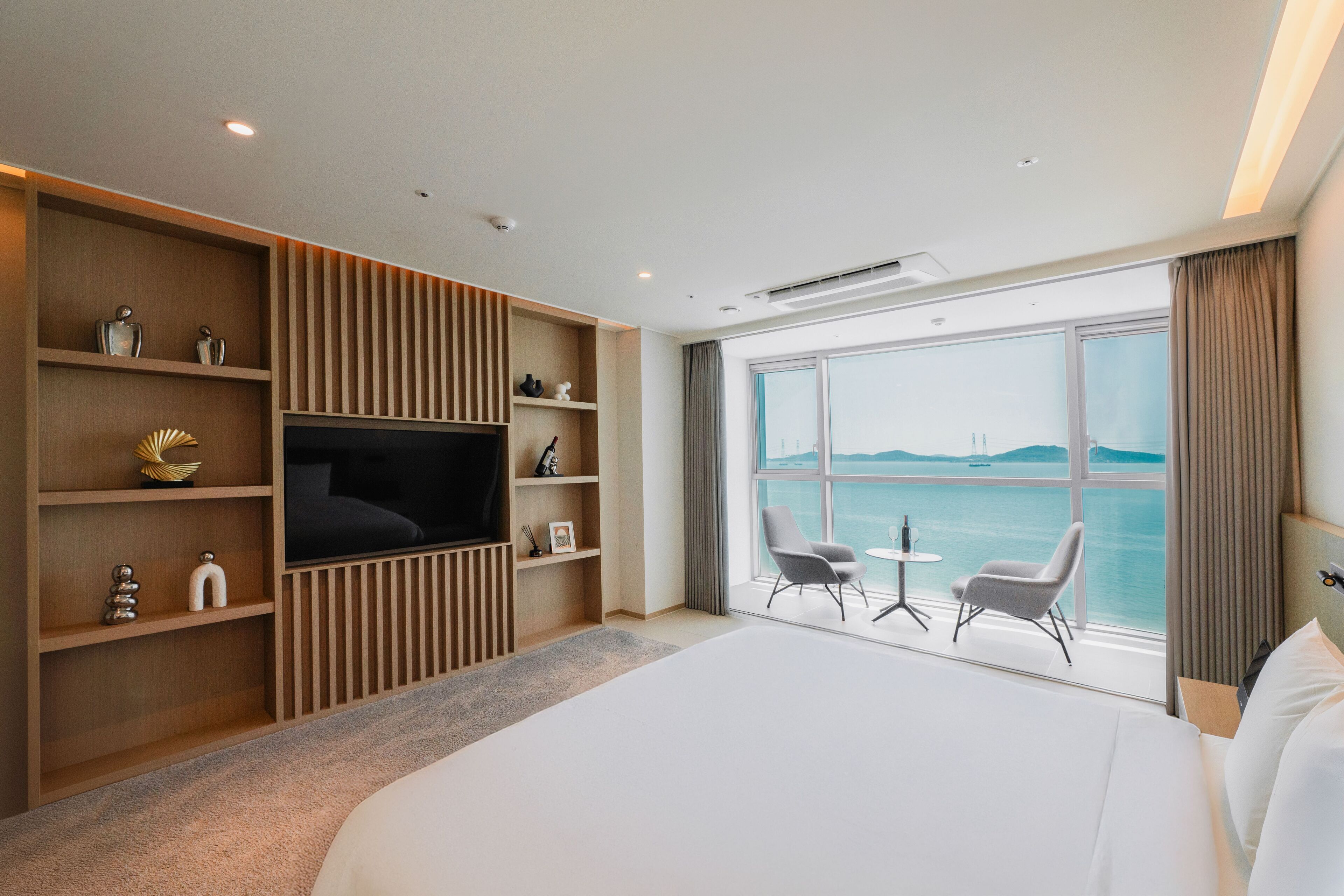 A hotel room with a large bed, a TV, a view of the sea, and a seating area with two chairs and a table.