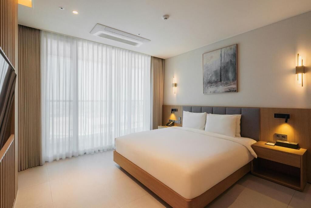 A modern hotel room with a large bed, bedside lamps, a painting on the wall, and a window with sheer curtains.