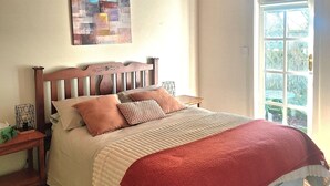 A bedroom with a wooden headboard, a bed with multiple pillows, a red blanket, a nightstand with a lamp, and a window with a view of greenery.