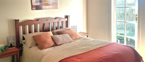 A bedroom with a wooden headboard, a bed with multiple pillows, a red blanket, a nightstand with a lamp, and a window with a view of greenery.