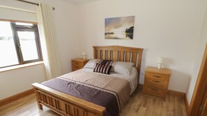 Un dormitorio con una cama de madera, dos mesitas de noche, una mesilla auxiliar, una ventana con cortinas y un cuadro en la pared.