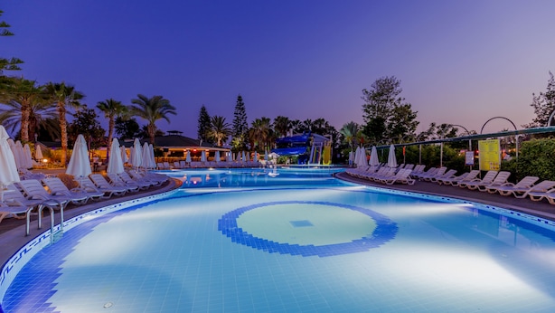 A swimming pool with lounge chairs and umbrellas, surrounded by palm trees and a building in the background. - Holiday Park Resort - All inclusive (Alanya)