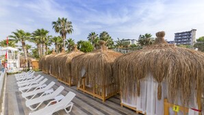 A row of thatched-roof huts with white curtains, wooden benches, and white lounge chairs on a tiled deck. - Holiday Park Resort - All inclusive (Alanya)