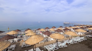 A beach with rows of white lounge chairs and straw umbrellas, a pier extending into the sea, and a flag on the left. - Holiday Park Resort - All inclusive (Alanya)