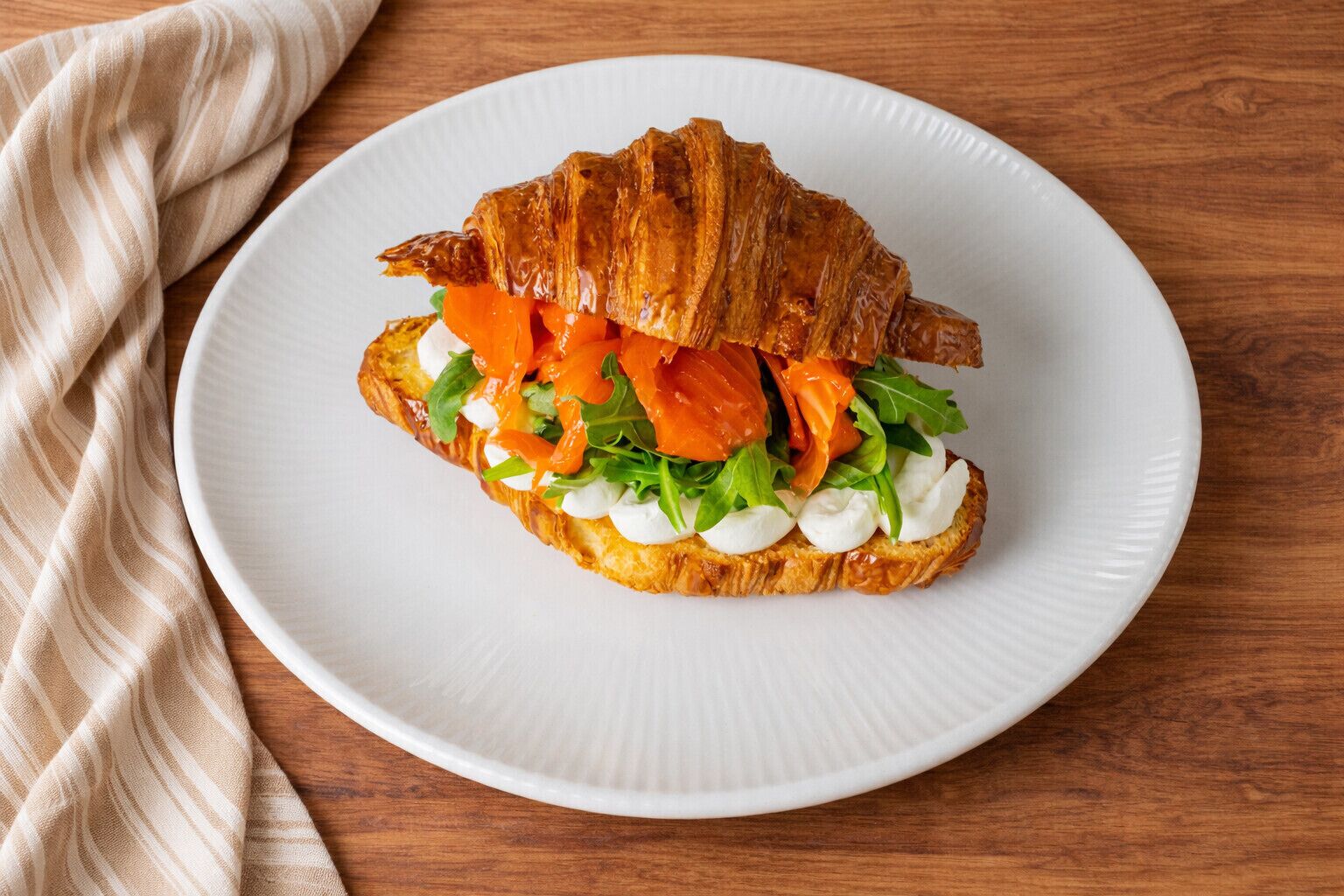 Sándwich de croissant con salmón ahumado, rúcula y queso crema sobre un plato blanco.