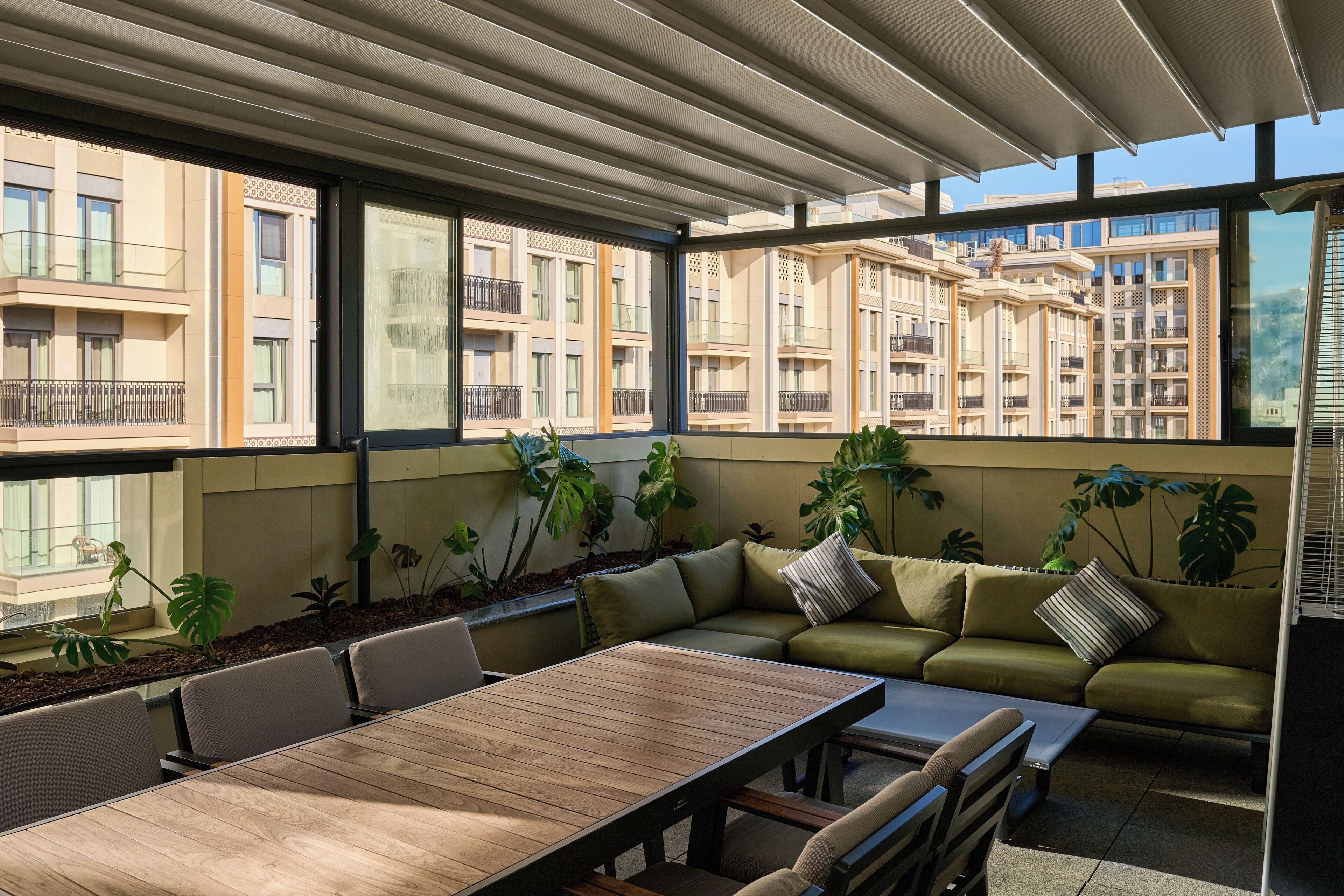 A modern outdoor seating area with a wooden table and cushioned seating, surrounded by large windows offering views of a cityscape.