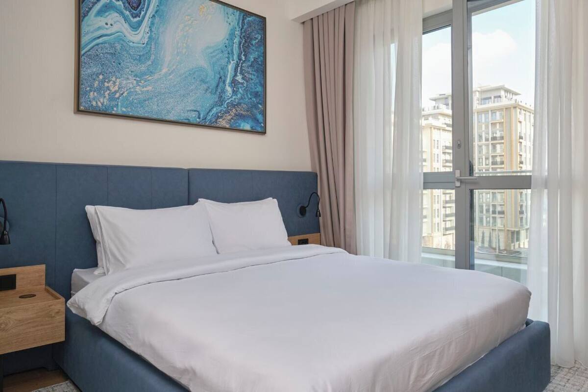 Une chambre d’hôtel moderne avec un grand lit, une table de chevet, une lampe fixée au mur et une vue sur la ville grâce à une grande fenêtre.