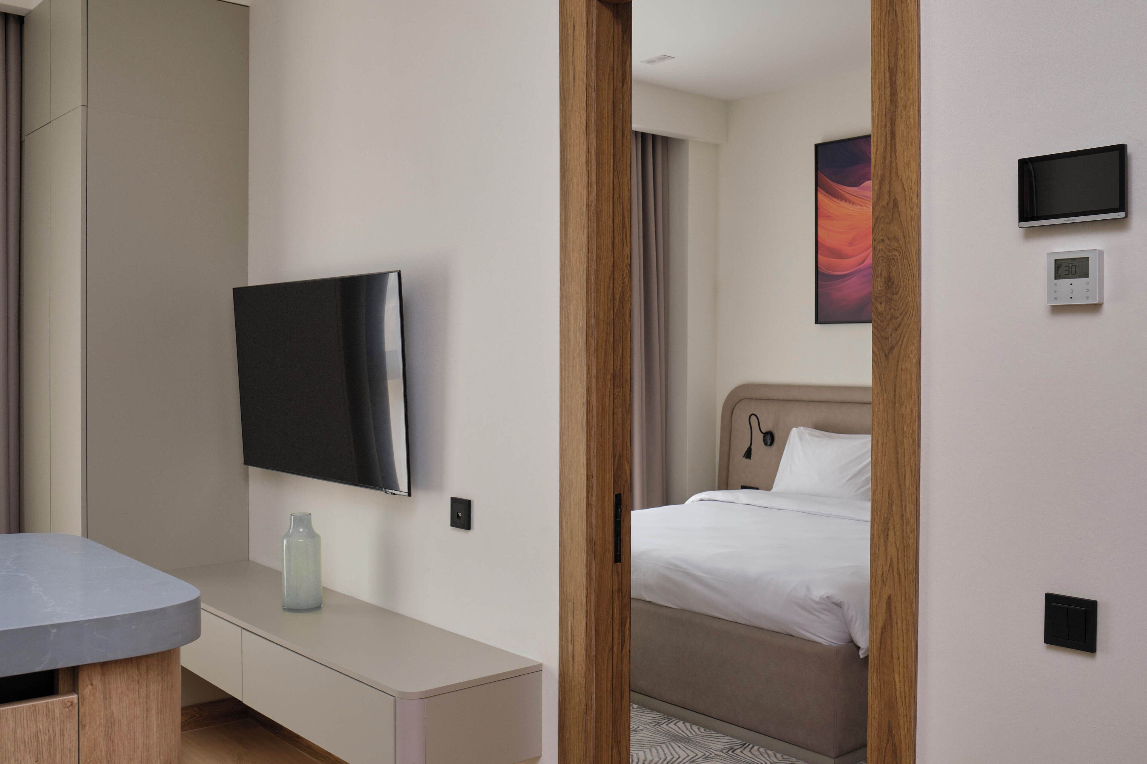 A modern hotel room with a flat-screen TV mounted on the wall, a bedside table with a vase, a bed with white bedding, and a wooden door frame.