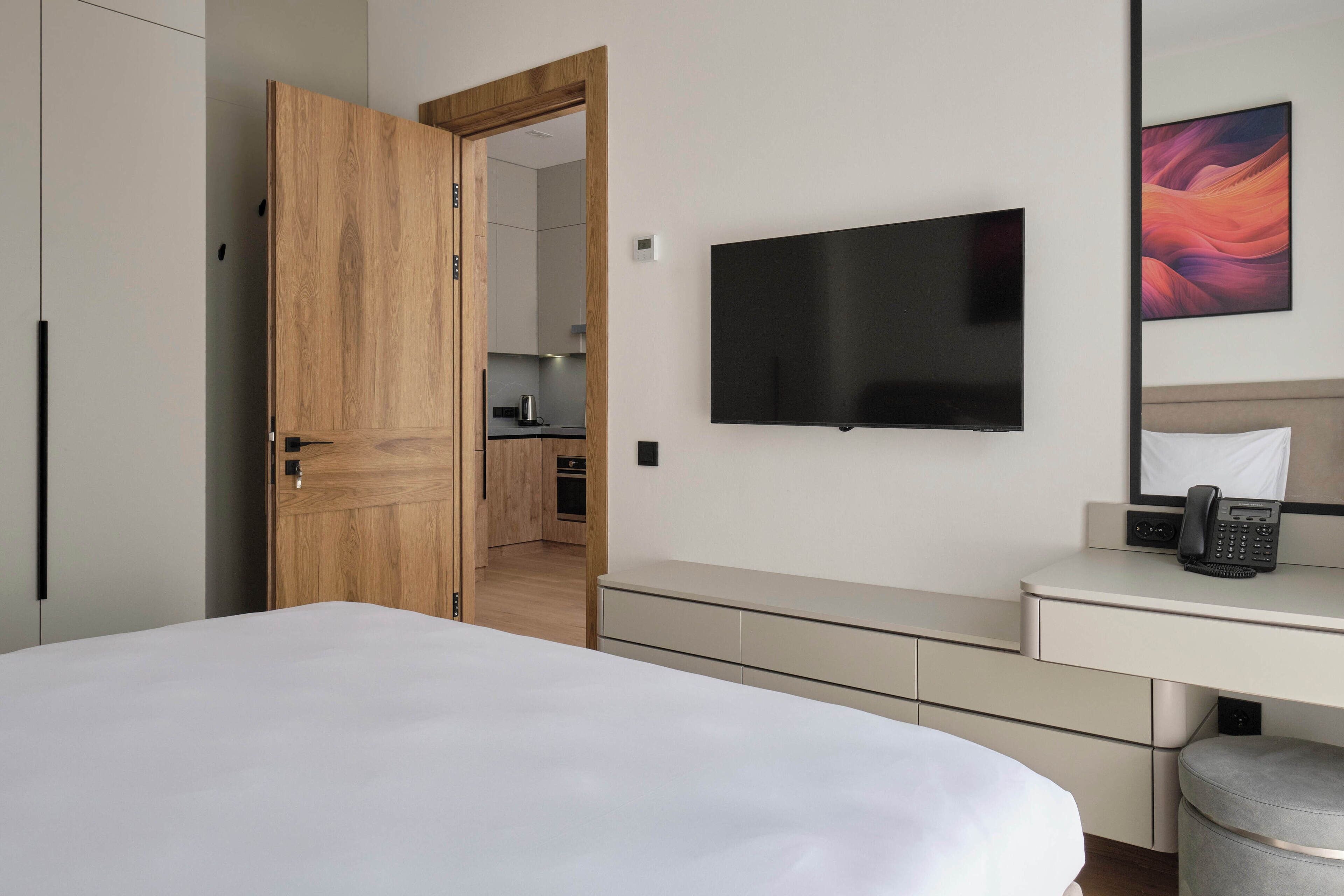 A hotel room with a bed, a desk with a telephone, a flat-screen TV, and a wooden door leading to a kitchenette.