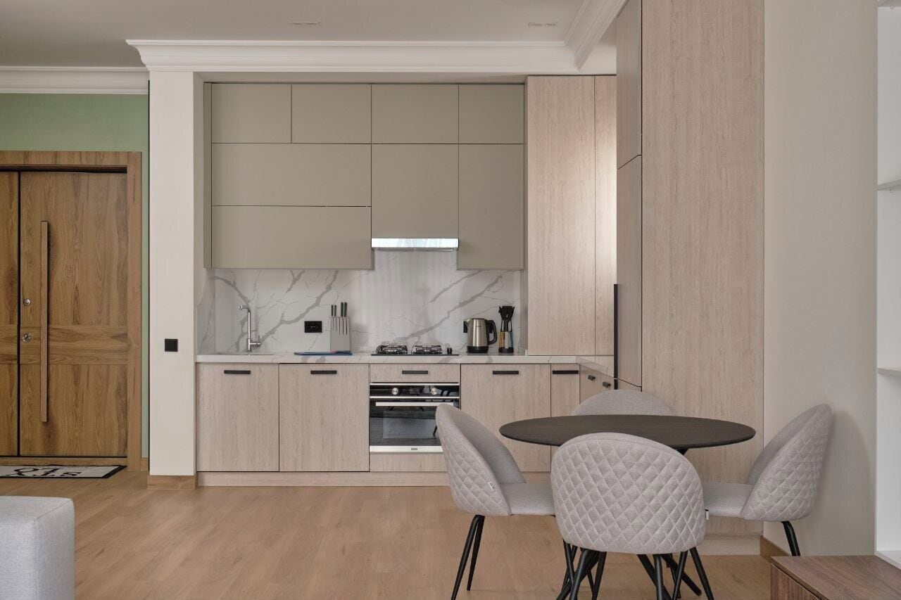 A modern kitchen with light wood cabinets, a marble backsplash, and a built-in oven. There is a round dining table with two chairs.