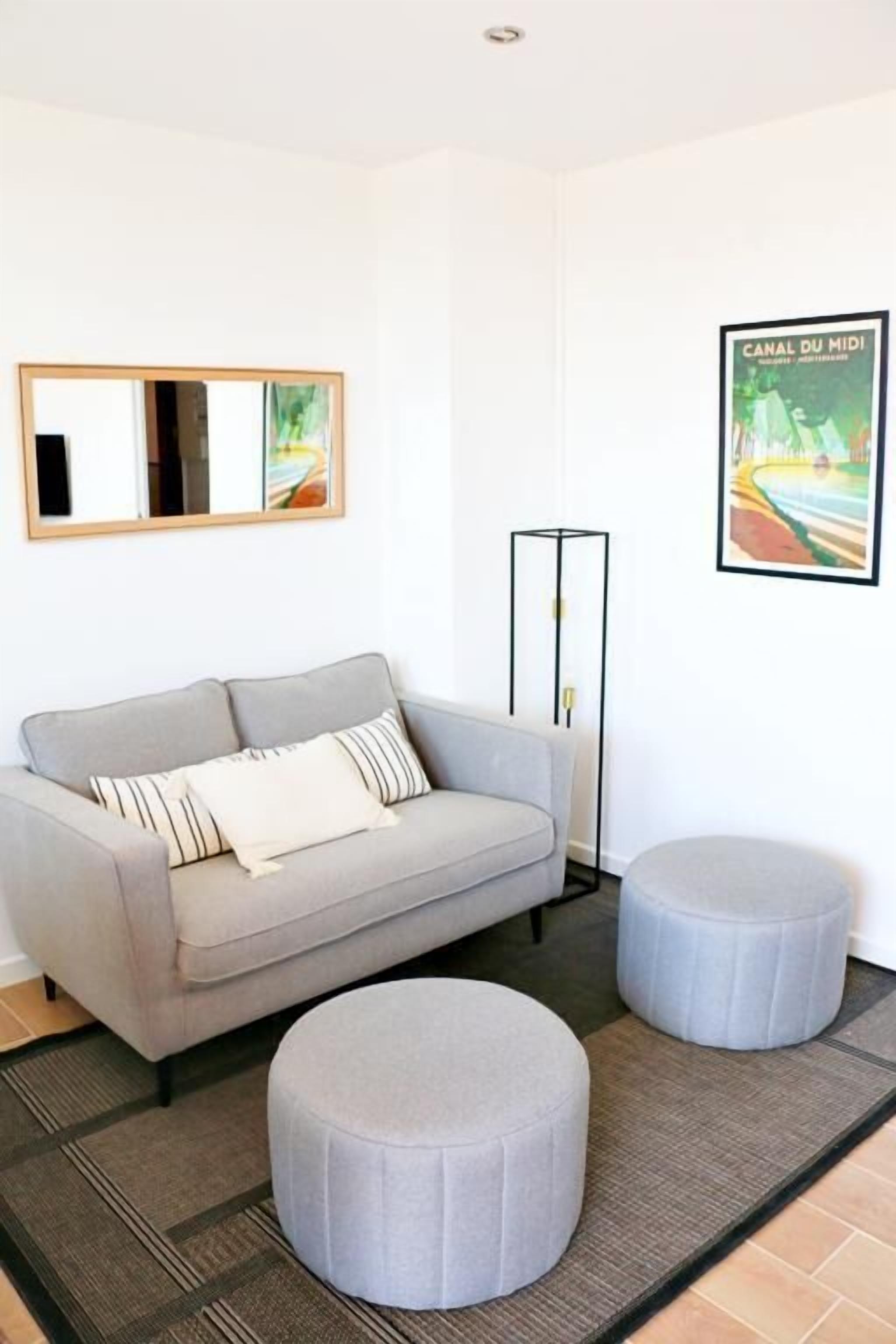 A modern living room with a grey sofa, two grey ottomans, a black floor lamp, and a framed picture of a canal.