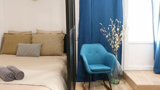 Een slaapkamer met een bed, kussens, een blauwe stoel en een vaas met takken.