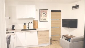 Een kleine keuken met witte kastjes, een zwart aanrecht en een houten deur. Een ingelijst schilderij hangt aan de muur, en een flatscreen televisie is boven het aanrecht bevestigd.