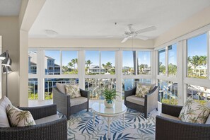 A spacious balcony with wicker furniture, a ceiling fan, and a view of a pool and palm trees.