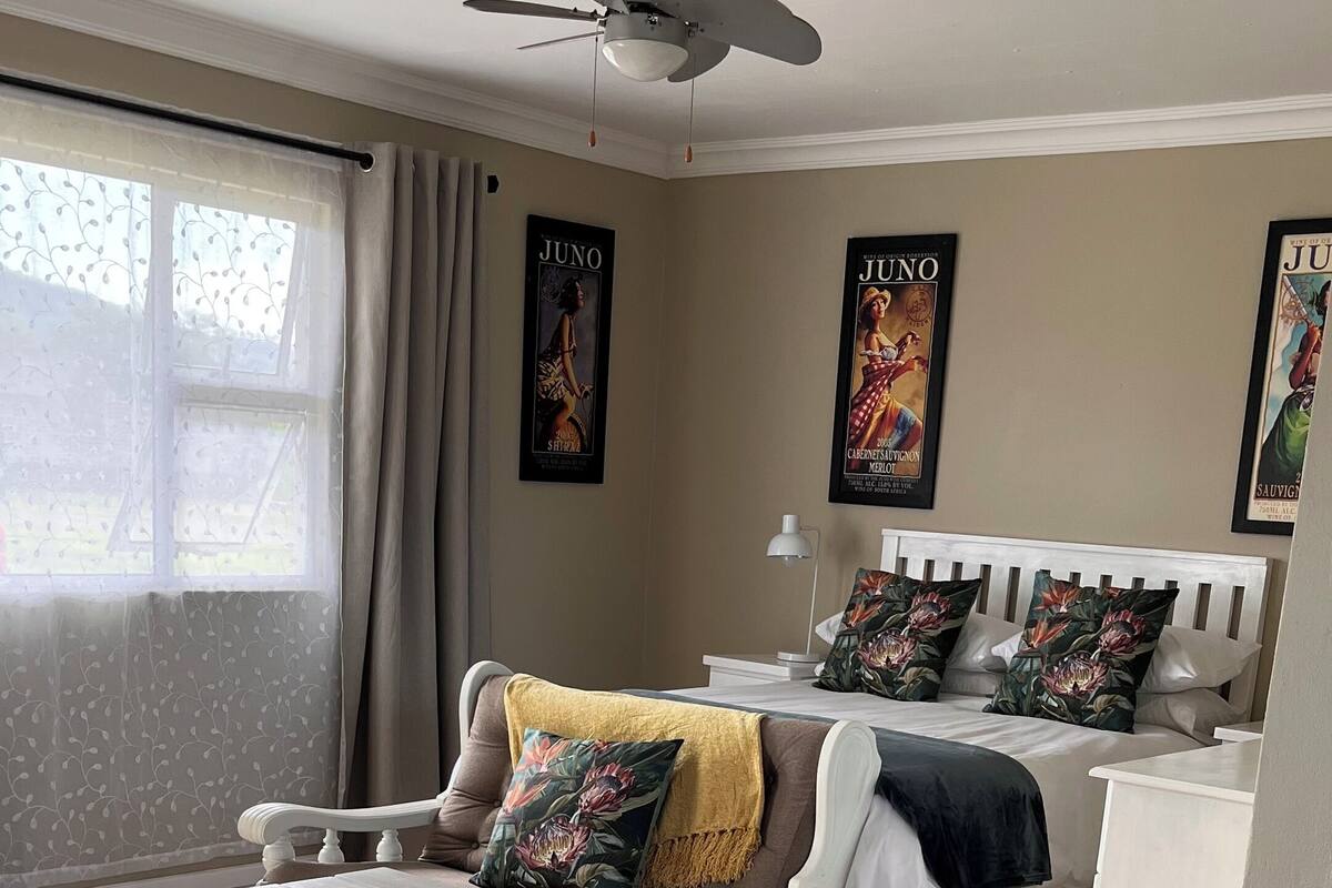 A room with a white table, a bench, a bed with floral pillows, a ceiling fan, and two posters on the wall.