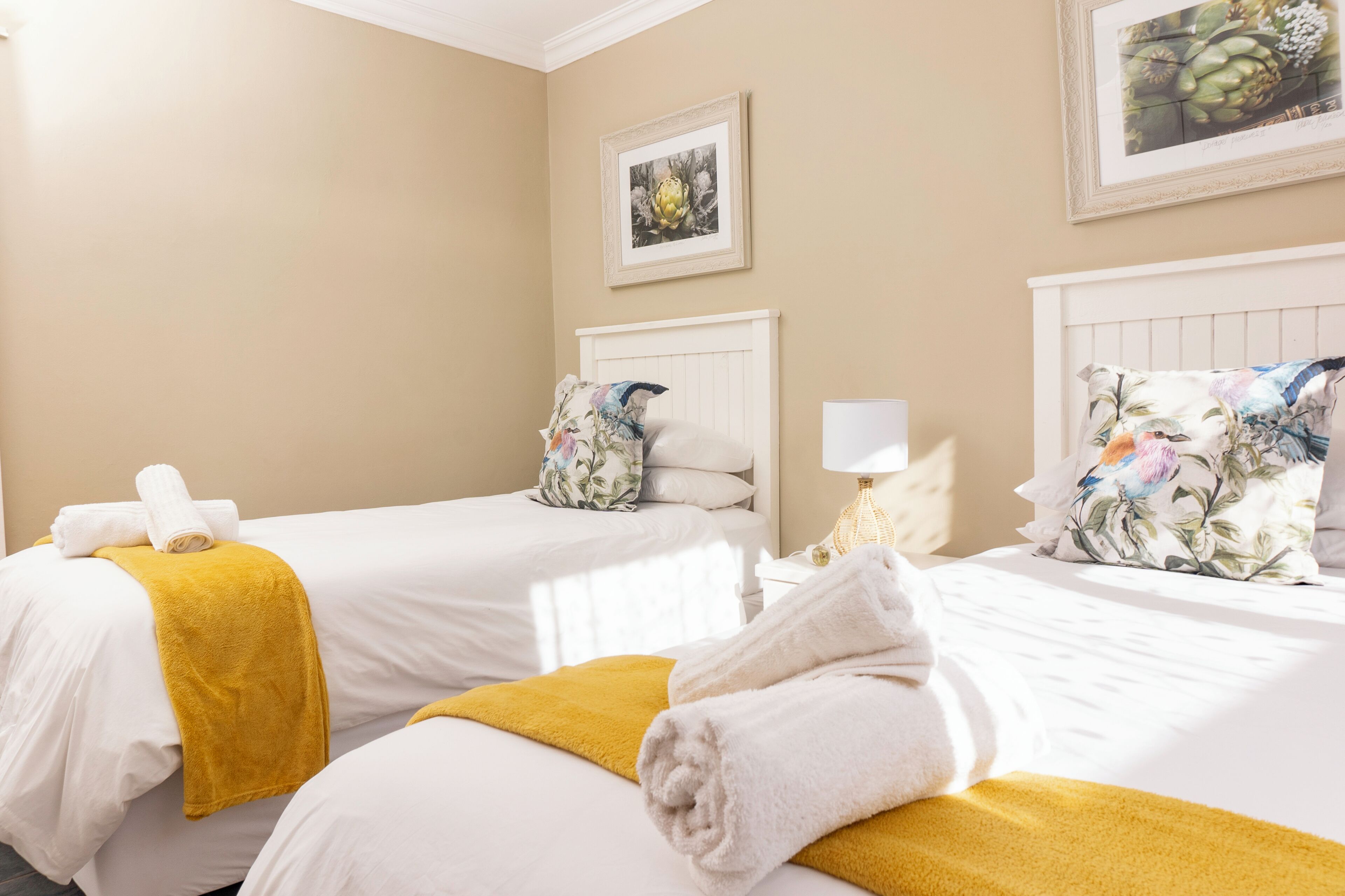 A hotel room with two beds, each with white linens and a yellow throw blanket. There is a white nightstand with a lamp and a framed picture on the wall.