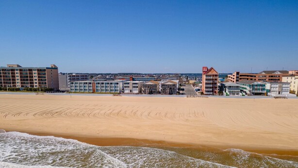 A beach with a sandy shore and waves. In the background, there are buildings with multiple floors and a red-roofed structure. - 1911 Oceanblock Summa (Ocean City)