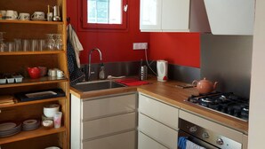 Una cocina compacta con armarios blancos, una pared roja, un estante de madera con vajilla, una ventana, una estufa y un horno.