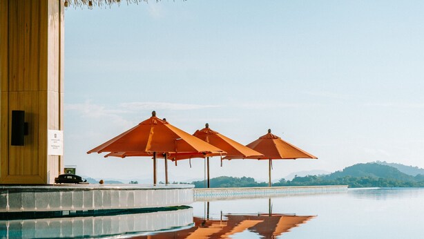 A pool area with three orange umbrellas, a wooden building, and a clear view of a distant mountain range. - Hilltop Wellness Living (Ratsada)