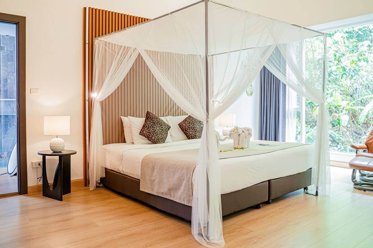 A modern bedroom with a four-poster bed, a bedside table with a lamp, and a view of greenery outside.
