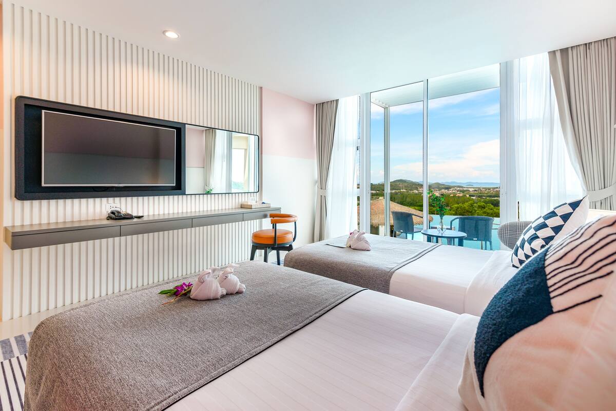 A hotel room with two beds, a TV mounted on the wall, a small table, and a view of the outdoors.
