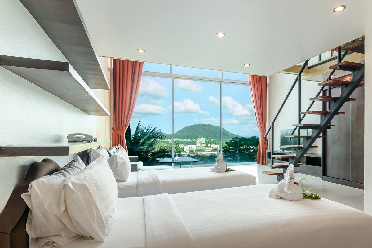 A modern hotel room with a large bed, a sofa, a dining table, and a staircase.