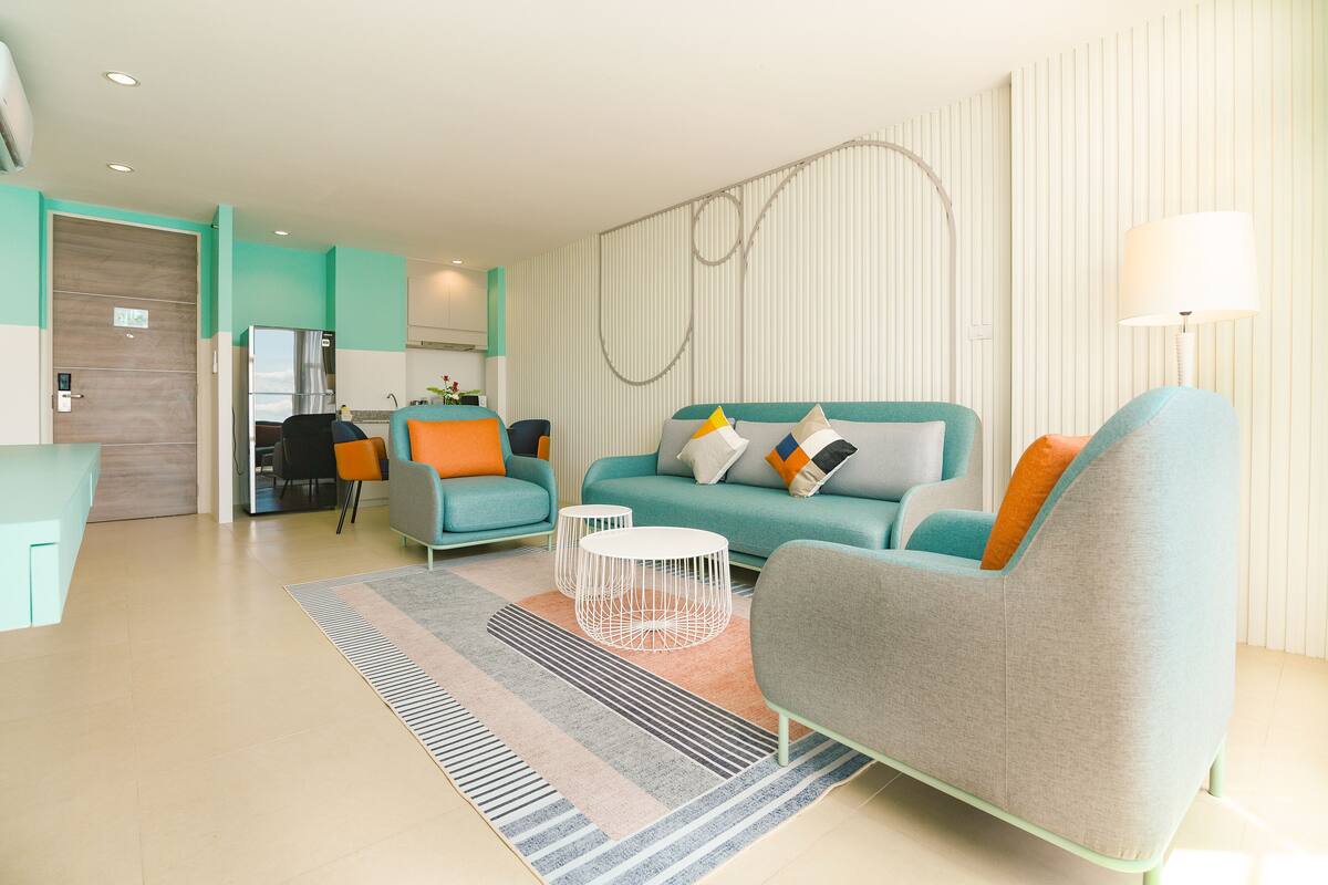 A modern living room with a teal sofa, grey armchair, and a small coffee table. A kitchen is visible in the background.