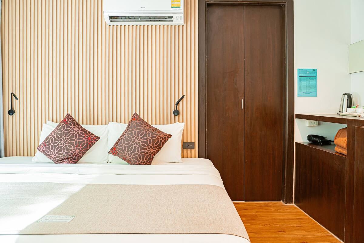 A hotel room with a bed, two patterned pillows, a wooden headboard, a wardrobe, and a wall-mounted air conditioner.