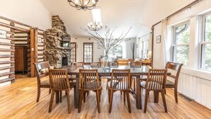 Comedor con chimenea de piedra, sillas de madera, candelabro y ventanas amplias.