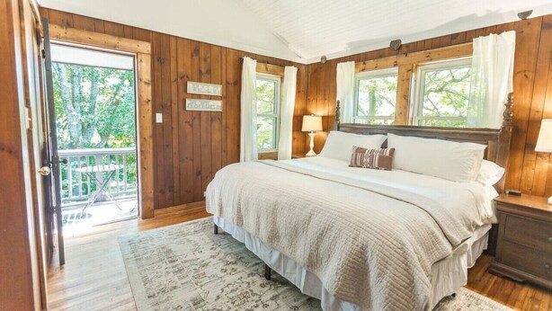 Un dormitorio con pared de paneles de madera, una cama con ropa de cama blanca, dos lámparas de noche y vistas a través de una ventana.